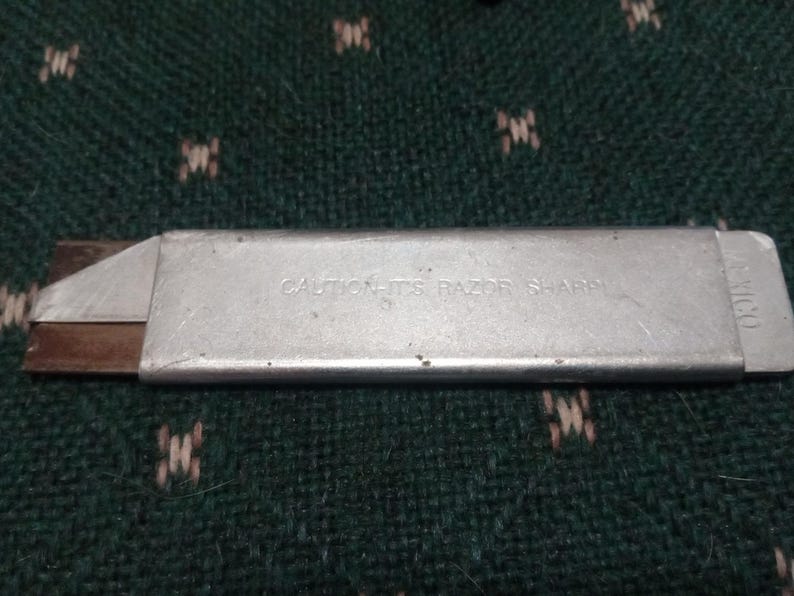 Op de afbeelding: Een zilveren hobbymes met een zichtbaar mes en de tekst "CAUTION-ITS RAZOR SHARP". Het mes ligt op een groen oppervlak met een patroon. Het uiteinde van het mes heeft het woord "MEXICO".