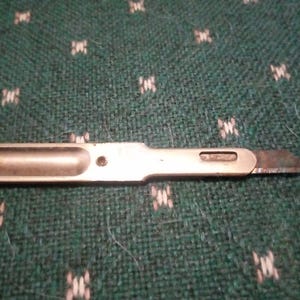 Op de afbeelding: Een close-up van een metalen scalpel met een stompe, schuine kling. De handgreep heeft een aangetaste zilverkleur en het lemmet vertoont tekenen van slijtage. De scalpel ligt op een groene stof met een patroon.