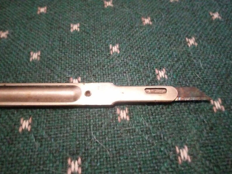 Op de afbeelding: Een close-up van een metalen scalpel met een stompe, schuine kling. De handgreep heeft een aangetaste zilverkleur en het lemmet vertoont tekenen van slijtage. De scalpel ligt op een groene stof met een patroon.