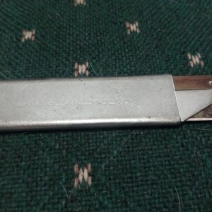 Op de afbeelding: Een zilveren hobbymes met een intrekbaar mes. Het mes is gedeeltelijk uitgeschoven en toont een scherpe rand. De tekst "CAUTION-ITS RAZOR SHARP" is zichtbaar op de metalen behuizing. Het mes ligt op een groen oppervlak met patroon.