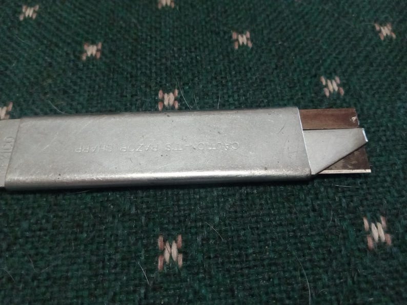 Op de afbeelding: Een zilveren hobbymes met een intrekbaar mes. Het mes is gedeeltelijk uitgeschoven en toont een scherpe rand. De tekst "CAUTION-ITS RAZOR SHARP" is zichtbaar op de metalen behuizing. Het mes ligt op een groen oppervlak met patroon.