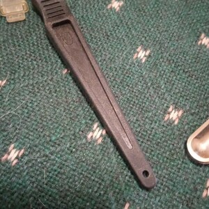 Op de afbeelding: Een zwart hobbymes met een zilveren mes en een getextureerd handvat. Het mes ligt op een groen oppervlak met een patroon. Een kleine, doorzichtige plastic hoes is zichtbaar linksboven.