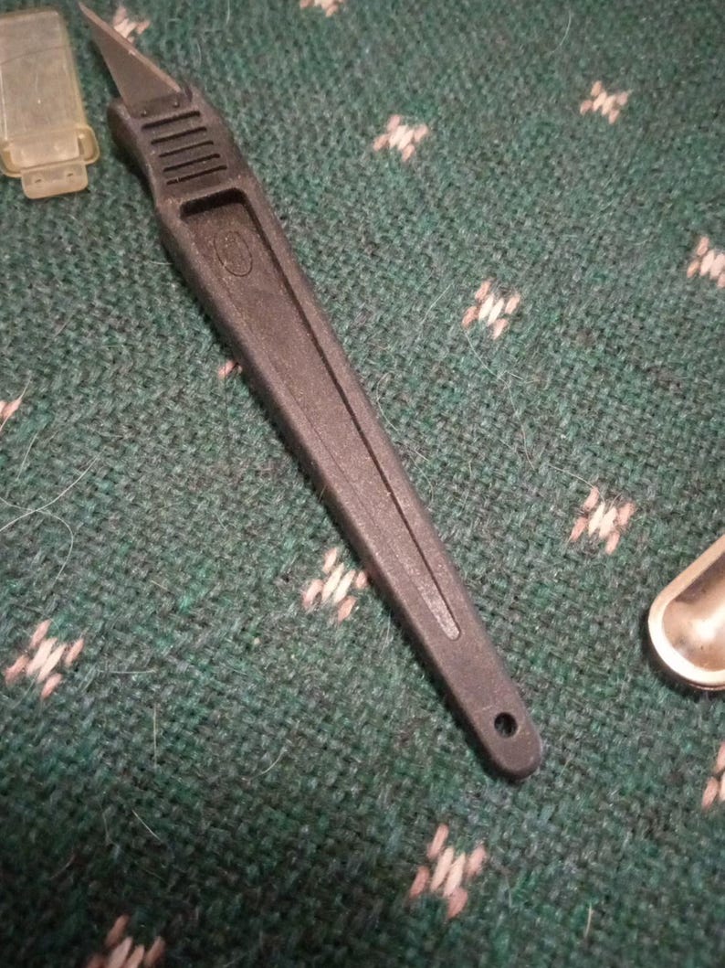 Op de afbeelding: Een zwart hobbymes met een zilveren mes en een getextureerd handvat. Het mes ligt op een groen oppervlak met een patroon. Een kleine, doorzichtige plastic hoes is zichtbaar linksboven.