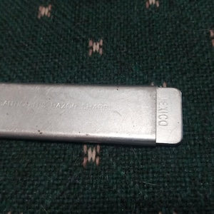 Op de afbeelding: Een close-up van een zilveren metalen object met de tekst "CAUTION IT'S RAZOR SHARP" en "MEXICO" erop gedrukt. Het object is rechthoekig en lijkt een mesbeschermer te zijn.