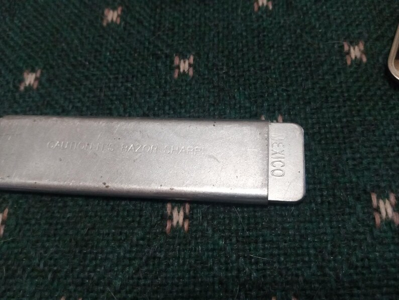 Op de afbeelding: Een close-up van een zilveren metalen object met de tekst "CAUTION IT'S RAZOR SHARP" en "MEXICO" erop gedrukt. Het object is rechthoekig en lijkt een mesbeschermer te zijn.