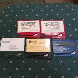 May include: A collection of business card holders in various colors. Two red holders with "Rocky Mountain Oldsmobile Club" text, a green "Edward Jones" holder, a black "Variety" holder, and a blue "Boys Town" holder with a ruler.