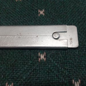 Op de afbeelding: Close-up van een zilveren metalen gereedschap met de tekst "TO OPEN AND CLOSE PRESS..." ge&euml;tst op het oppervlak. Een kleine, cirkelvormige klinknagel is zichtbaar nabij het uiteinde. Het gereedschap is rechthoekig en lijkt voor een specifiek doel te worden gebruikt.