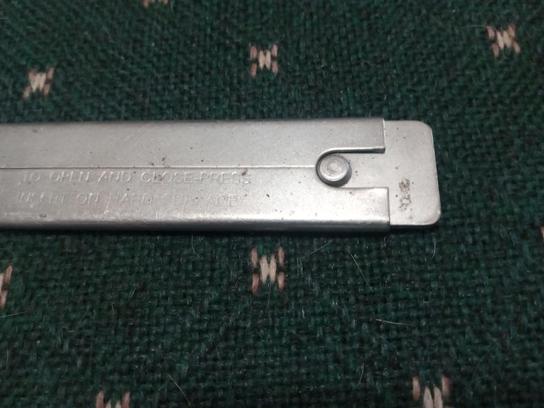 Op de afbeelding: Close-up van een zilveren metalen gereedschap met de tekst "TO OPEN AND CLOSE PRESS..." ge&euml;tst op het oppervlak. Een kleine, cirkelvormige klinknagel is zichtbaar nabij het uiteinde. Het gereedschap is rechthoekig en lijkt voor een specifiek doel te worden gebruikt.