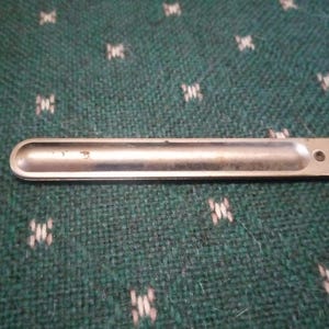 Op de afbeelding: Een zilverkleurige metalen spatel met een afgerond uiteinde en een klein gaatje bij het handvat. De spatel is ontworpen voor laboratorium- of keukengebruik. De achtergrond is een groene stof met een patroon.