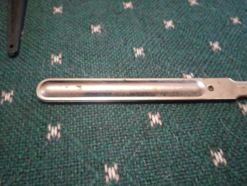 Op de afbeelding: Een zilverkleurige metalen spatel met een afgerond uiteinde en een klein gaatje bij het handvat. De spatel is ontworpen voor laboratorium- of keukengebruik. De achtergrond is een groene stof met een patroon.