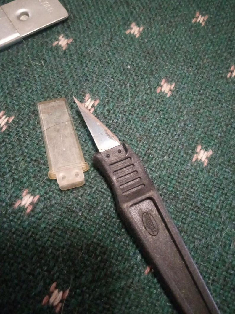 Op de afbeelding: Een zwart hobbymes met een zilveren mes en een doorzichtige plastic mesbeschermer. Het mes ligt op een groen oppervlak met een patroon. Het mes is blootgesteld en klaar voor gebruik.