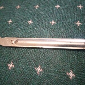 Op de afbeelding: Een zilverkleurig metalen gereedschap met een afgerond uiteinde en een klein, rechthoekig gedeelte aan de linkerkant. Het gereedschap ligt op een groene stof met een herhalend wit en lichtgroen patroon.