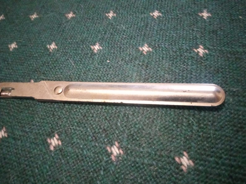 Op de afbeelding: Een zilverkleurig metalen gereedschap met een afgerond uiteinde en een klein, rechthoekig gedeelte aan de linkerkant. Het gereedschap ligt op een groene stof met een herhalend wit en lichtgroen patroon.