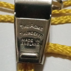 May include: Close-up of a silver metal whistle with the text "The ACME Thunderer" and "MADE IN ENGLAND" engraved on the front. A yellow cord is attached to the whistle.