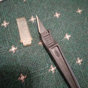 Op de afbeelding: Een zwart hobbymes met een zilveren mes en een doorzichtige plastic mesbeschermer. De handgreep is zwart en heeft een geribbeld ontwerp voor grip. Het mes is scherp en puntig.