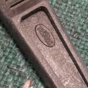 Op de afbeelding: Close-up van een zwarte plastic gereedschapshandgreep met de in reli&euml;f aangebrachte tekst "Hessiors" in een ovaal. De handgreep heeft een getextureerd oppervlak en verschillende parallelle groeven. De achtergrond is een groen en roze patroonstof.