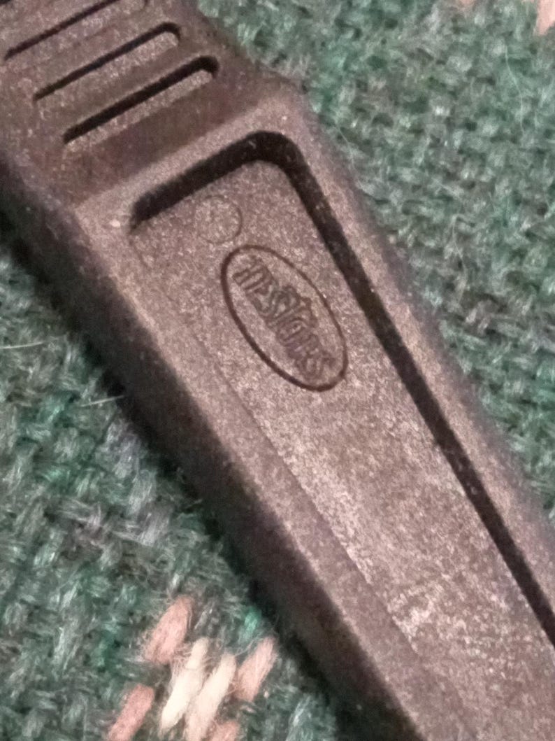 Op de afbeelding: Close-up van een zwarte plastic gereedschapshandgreep met de in reli&euml;f aangebrachte tekst "Hessiors" in een ovaal. De handgreep heeft een getextureerd oppervlak en verschillende parallelle groeven. De achtergrond is een groen en roze patroonstof.