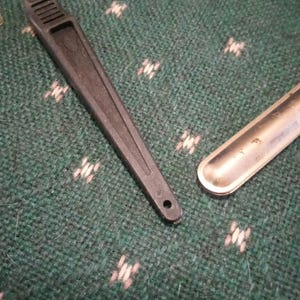 Op de afbeelding: Een zwarte plastic briefopener met een zilverkleurig mes en een klein gaatje aan het uiteinde van de handgreep. Daarnaast ligt een klein, zilverkleurig metalen voorwerp. De items liggen op een groen oppervlak met patroon.