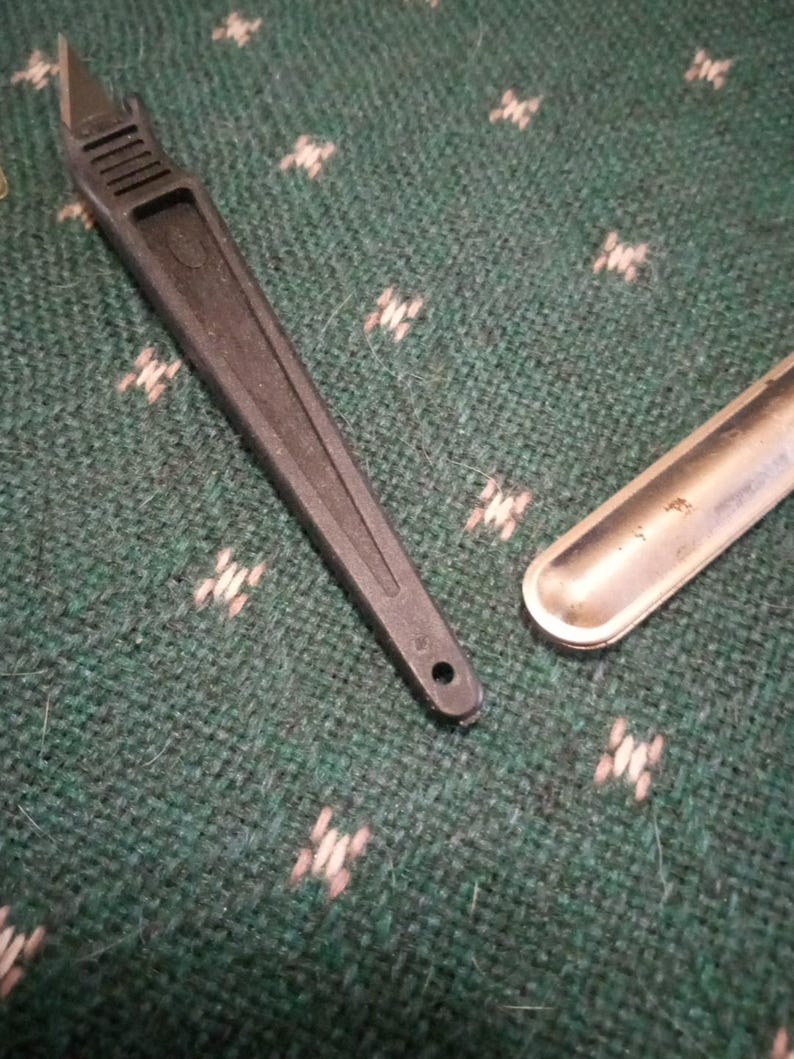 Op de afbeelding: Een zwarte plastic briefopener met een zilverkleurig mes en een klein gaatje aan het uiteinde van de handgreep. Daarnaast ligt een klein, zilverkleurig metalen voorwerp. De items liggen op een groen oppervlak met patroon.