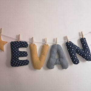 Könnte beinhalten: Ein Kinderzimmerbanner mit dem Namen "EVAN", der in Stoffbuchstaben geschrieben ist. Die Buchstaben sind mit Ankern, Sternen und Streifen verziert. An jedem Ende des Banners hängt ein gelber Stern und ein Halbmond.