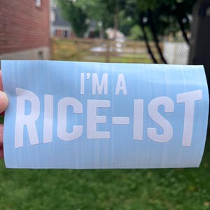 May include: A light blue rectangular vinyl decal featuring the white text I'M A RICE-IST in a bold, sans-serif font. The decal has a smooth, matte finish and is presented on a light blue backing sheet, displaying clear, legible lettering.
