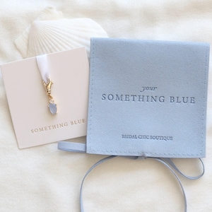 Può includere: Un astuccio e un biglietto azzurri con la scritta "SOMETHING BLUE" e un ciondolo dorato con una gemma blu. L'astuccio riporta anche la scritta "your SOMETHING BLUE" e "BRIDAL CHIC BOUTIQUE". Gli oggetti sono su una superficie bianca con una conchiglia.