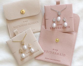 May include: Two pink velvet jewelry boxes with gold clasps. The boxes have the text "to my bridesmaid" and "to my matron of honor" printed on them. Each box contains a pearl necklace and earrings.