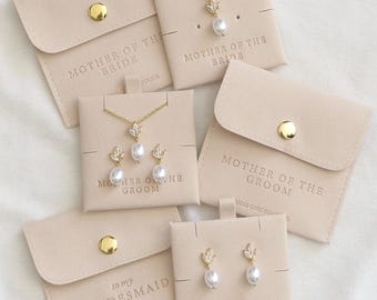 Könnte beinhalten: Goldfarbene Halsketten und Ohrringe mit weißen perlenartigen Tropfen und blattförmigen Akzenten auf beigen Karten. Die Karten sind mit Mother of the Bride, Mother of the Groom und Bridesmaid beschriftet und haben passende beige Druckknopfbeutel.