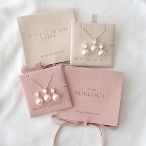 Könnte beinhalten: Brautschmuck-Sets mit Perlen- und Kristallhalsketten und Ohrringen, präsentiert in rosafarbenen und beigen Beuteln. Die Beutel sind mit Texten wie "Mother of the Bride" und "To my Bridesmaid" geprägt.