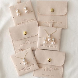 May include: A collection of jewelry sets, including earrings and necklaces, presented on light beige cards and pouches. Each set features pearls and clear gemstones. Text on the cards reads "to my Maid of Honour", "to my Bridesmaid", and "Mother of the Bride".