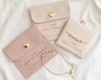 May include: Two pink velvet pouches with gold buttons and the text "to my bridesmaid" and "to my maid of honor" printed on them. Each pouch contains a delicate gold chain bracelet with small flower-shaped charms.