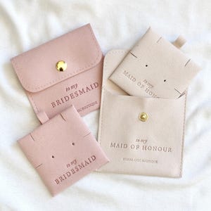 May include: Three pink and beige velvet pouches with gold buttons and the text "to my bridesmaid" and "to my maid of honor" printed on them.