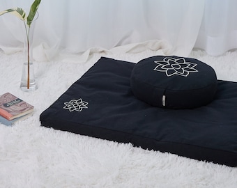 May include: A black meditation cushion set with a rectangular mat and a round cushion. Both pieces feature a white floral design. The set is placed on a white, fluffy rug, with a clear vase and a book visible in the background.