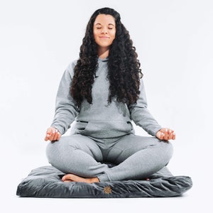 May include: A person sits in a meditative pose on a gray meditation cushion with a gold logo. The person is wearing a gray hoodie and gray sweatpants.