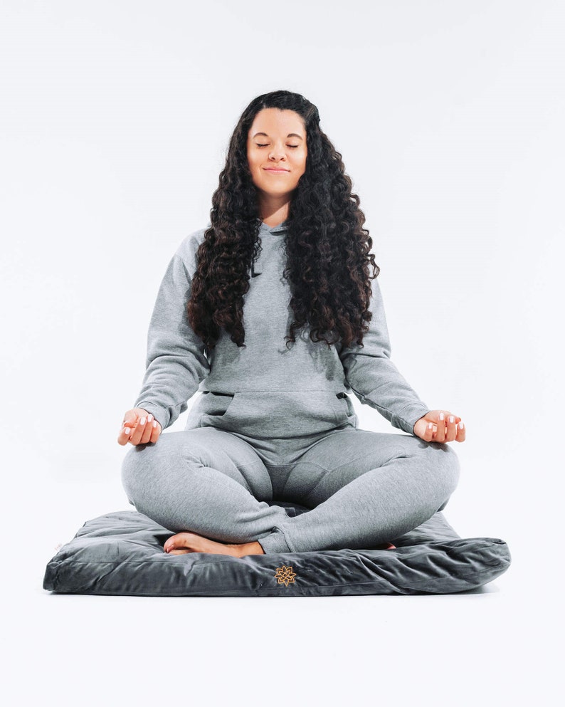 May include: A person sits in a meditative pose on a gray meditation cushion with a gold logo. The person is wearing a gray hoodie and gray sweatpants.