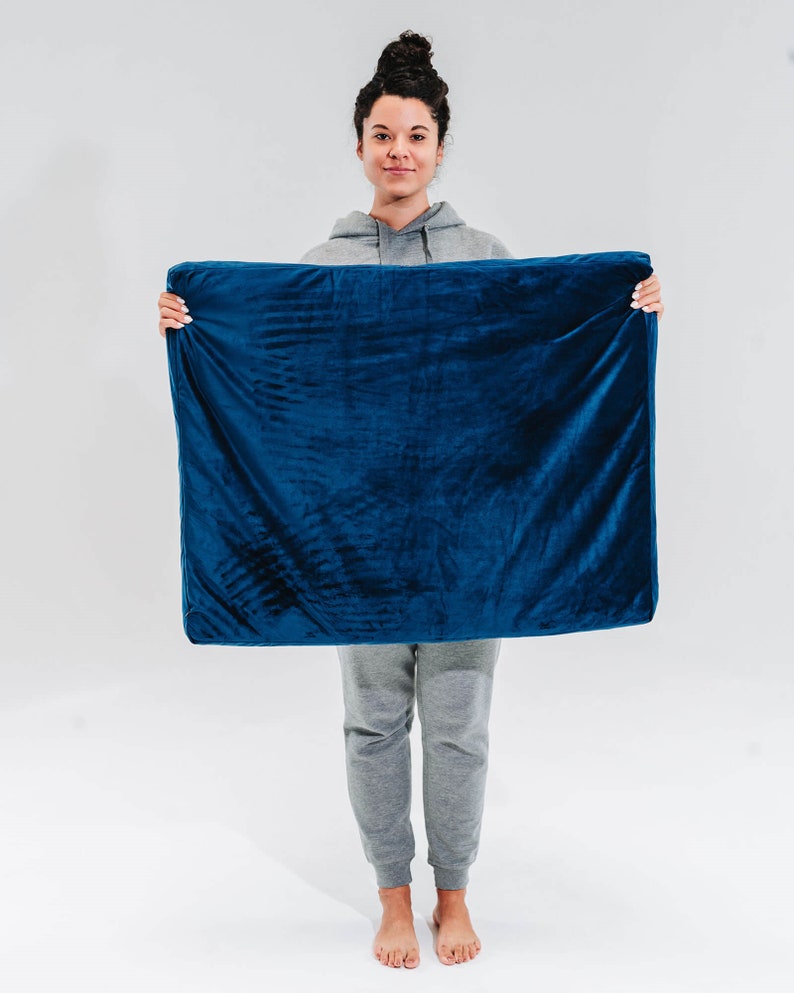 May include: A person holding a large, dark blue, rectangular cushion or pillow. The cushion is made of a soft, plush fabric.