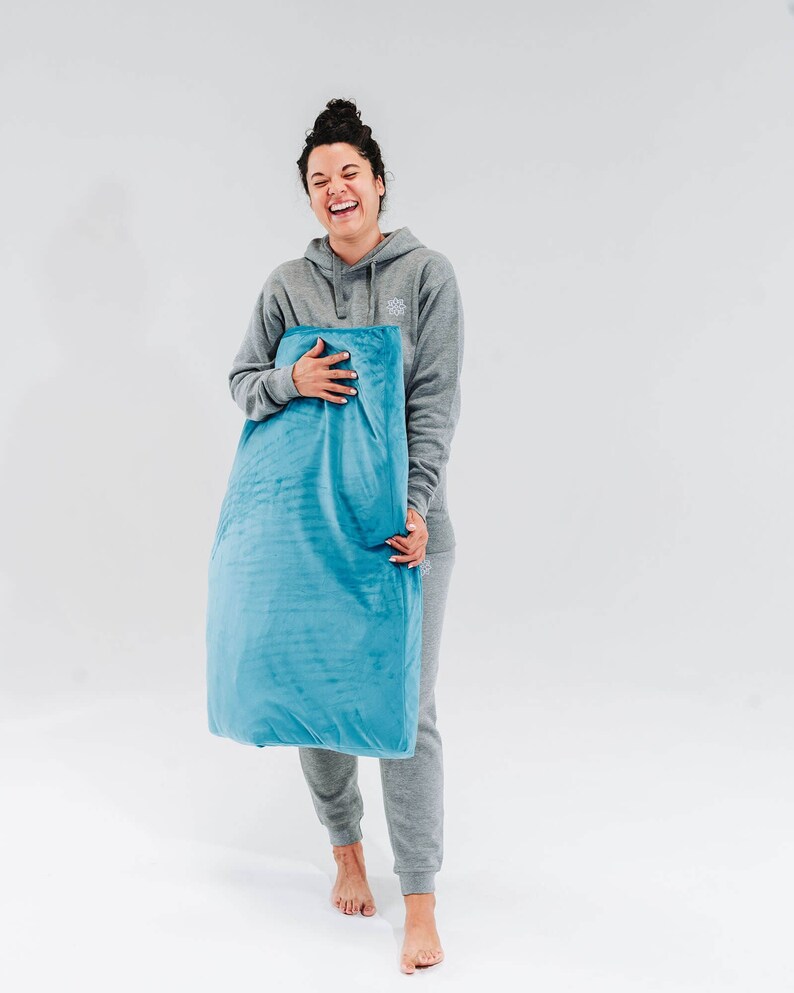 May include: A woman in a gray hoodie and matching sweatpants is holding a blue pillow. She is smiling and looking at the camera.