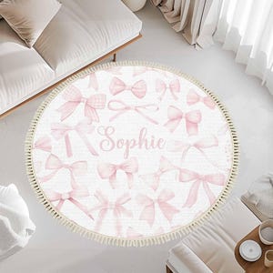 Può includere: Tappeto rotondo bianco con un motivo a fiocchi rosa e il nome "Sophie" al centro. Il tappeto ha un bordo con frange. I fiocchi sono in varie tonalità di rosa. Il tappeto è posizionato su un pavimento bianco.