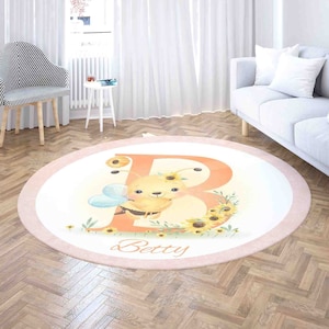May include: A round pink and white rug with a large orange letter B surrounded by yellow sunflowers and a cartoon bee with a blue and white wings. The name "Betty" is written below the letter.