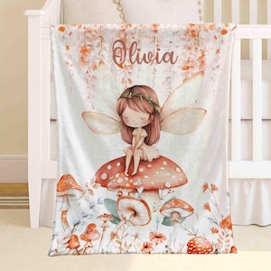 May include: A white baby blanket with a fairy illustration sitting on a mushroom. The blanket features a floral design and the name "Olivia". The color scheme includes orange, brown, and white. The blanket is designed for a cot or crib.