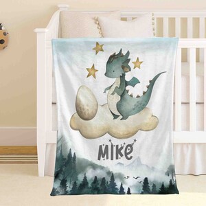 Puede incluir: Una manta de forro polar suave y blanca con un dragón verde y un huevo marrón en una nube. La manta tiene una escena de bosque en la parte inferior y el nombre "Mike" escrito en negro.