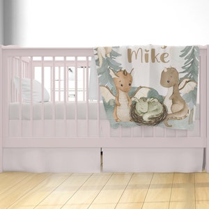 May include: A light pink cot with a white fitted sheet and a white pillow. A soft, green and brown baby blanket with a dragon family design and the name "Mike" is draped over the cot.