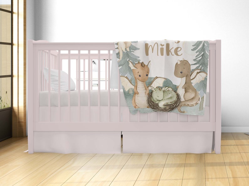 May include: A light pink cot with a white fitted sheet and a white pillow. A soft, green and brown baby blanket with a dragon family design and the name "Mike" is draped over the cot.