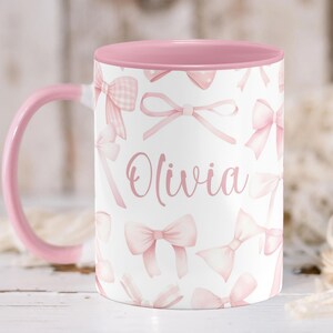 Pu&ograve; includere: Una tazza in ceramica rosa con sfondo bianco e bordo rosa. La tazza presenta un motivo a fiocco rosa e il nome "Olivia" scritto in lettere rosa.