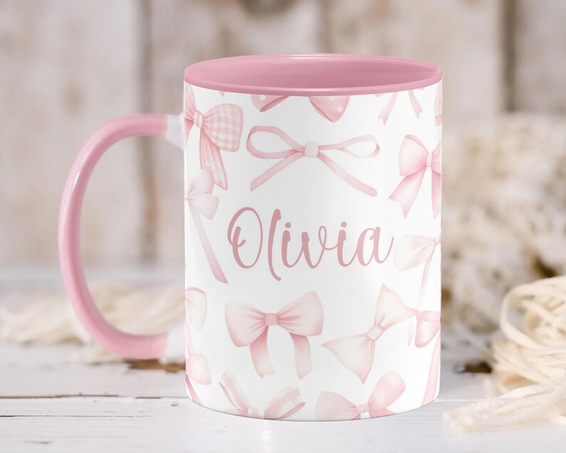 Pu&ograve; includere: Una tazza in ceramica rosa con sfondo bianco e bordo rosa. La tazza presenta un motivo a fiocco rosa e il nome "Olivia" scritto in lettere rosa.