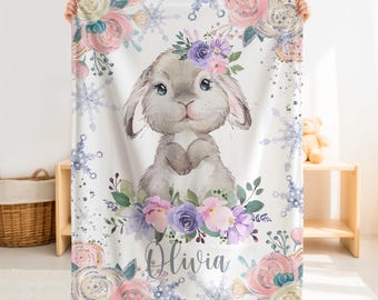 May include: A white fleece blanket featuring a central illustration of a grey bunny surrounded by purple and pink flowers and blue snowflakes. The name Olivia is printed in a cursive script below the bunny, framed by floral patterns.