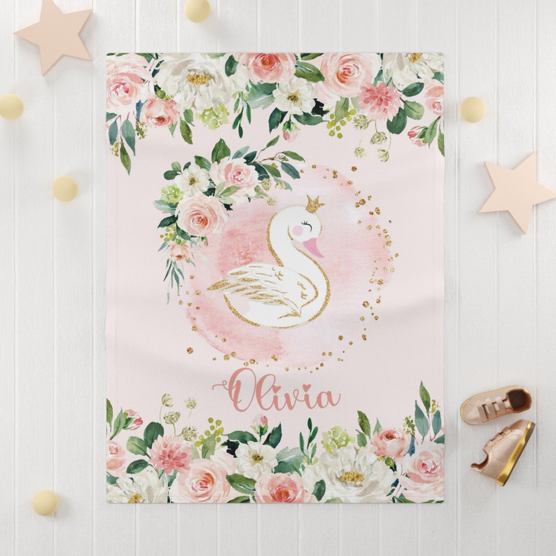 May include: A pink and white floral baby blanket with a gold glitter swan and the name "Olivia" written in cursive.