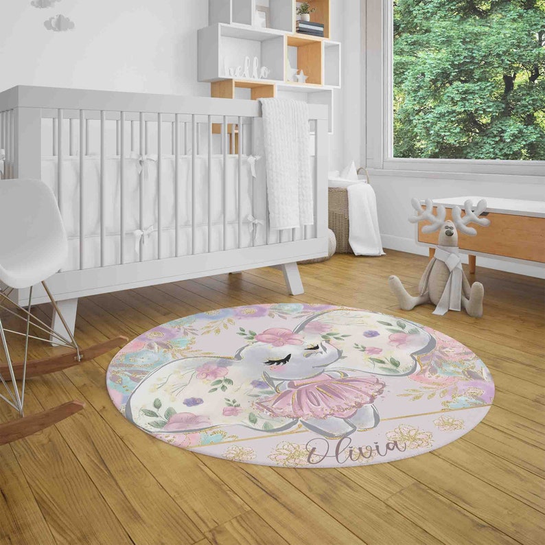 May include: A round pink and purple nursery rug with a floral design and a gray elephant wearing a pink tutu. The rug has the name "Olivia" written in gold glitter.