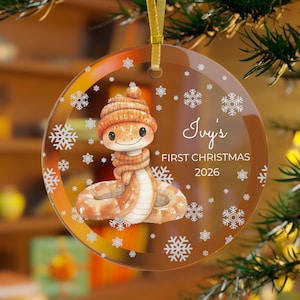 Op de afbeelding: Een ronde, doorzichtige acryl kerstornament met een cartoon slang die een beige gebreide muts en sjaal draagt. Het ontwerp bevat witte sneeuwvlokpatronen en de tekst Ivy's FIRST CHRISTMAS 2026. Aan de bovenkant zit een gouden lintje bevestigd.
