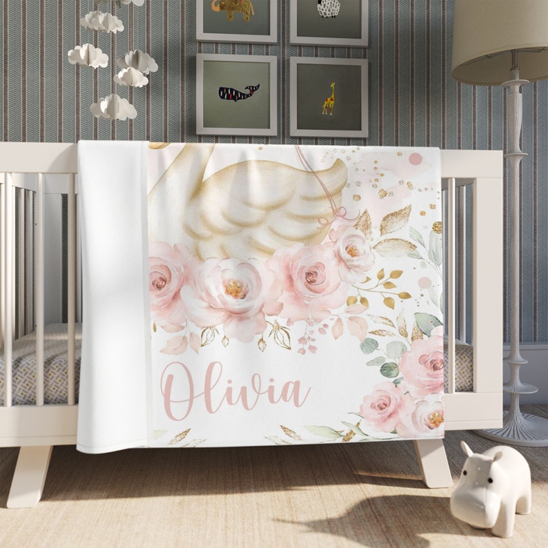 May include: A white baby blanket with a pink and gold floral design and a swan. The name Olivia is written in pink script.